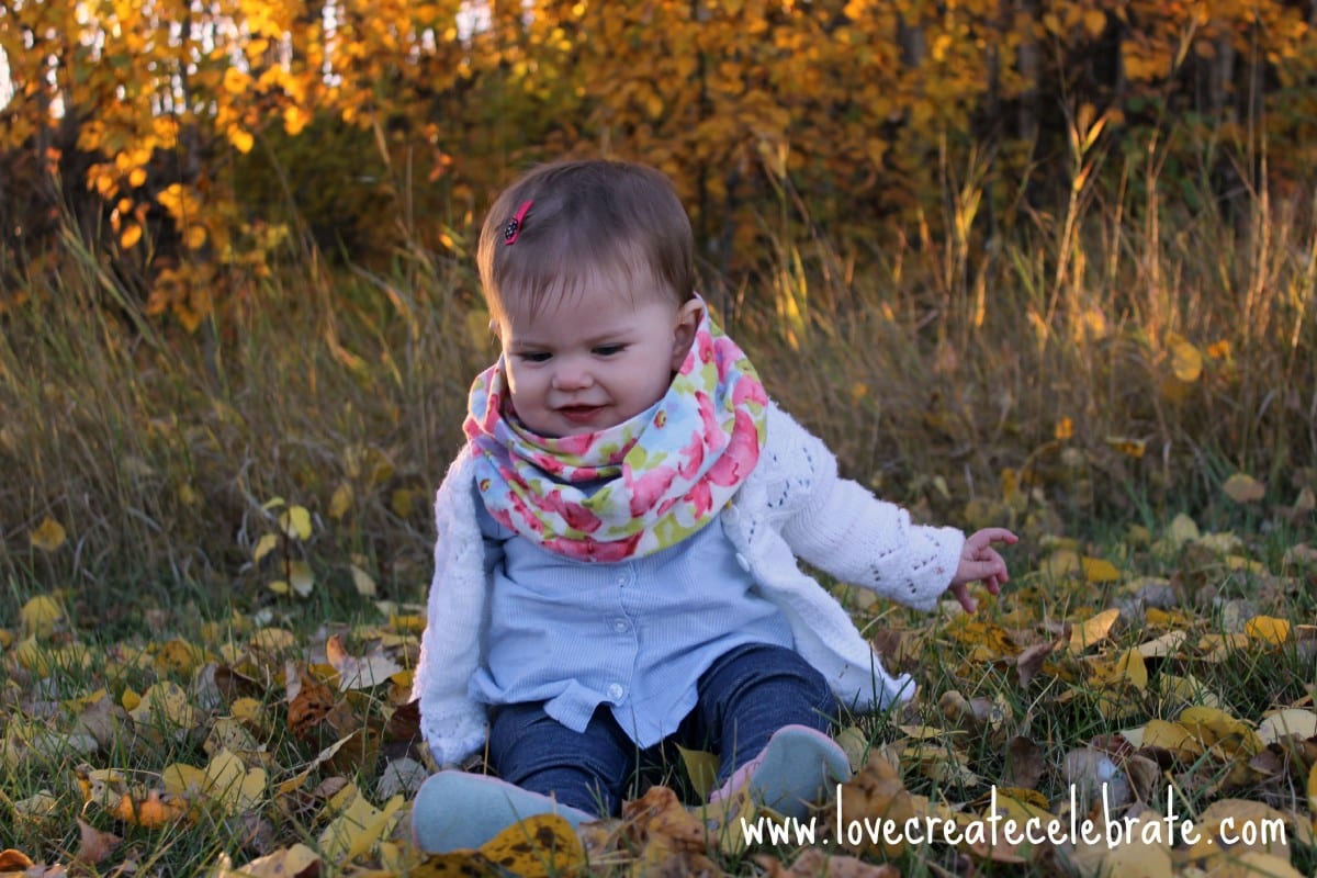 Baby girl sitting in the leaves on the ground with a flowered baby infinity scarf.