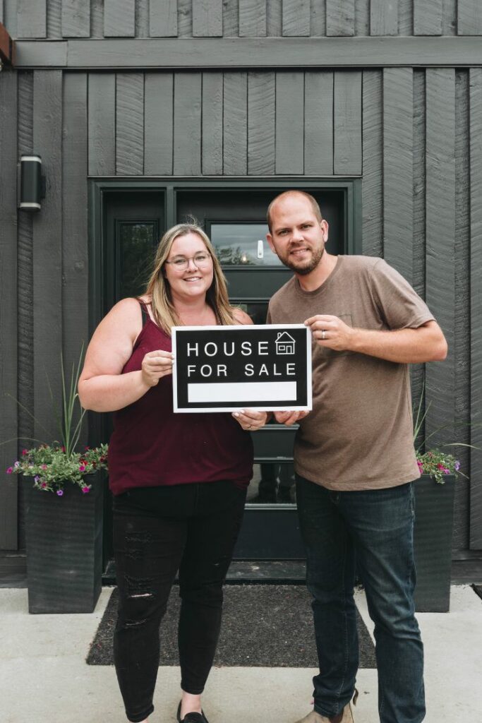 Couple holding a house for sale sign in front of their home